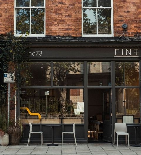 Exterior view of FINT, featuring a brick facade with large windows, a black storefront sign, and outdoor seating with white chairs.