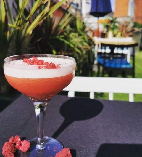 A refreshing cocktail with a frothy top and garnished with red berries, presented on a table at H A R V E Y • N I C H O L S • Fourth Floor • Brasserie & Bar, with a sunny outdoor seating area in the background.