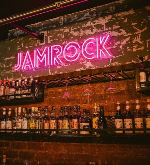 Neon sign reading "JAMROCK" above a bar shelf with assorted bottles of liquor, set against a rustic wall with peeling paint, creating an ambient nightlife atmosphere.