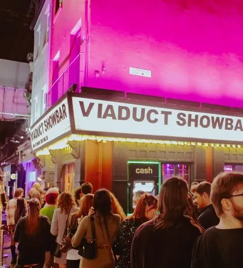 Crowd of people outside Viaduct Showbar at night with vibrant pink lighting on the building's facade.