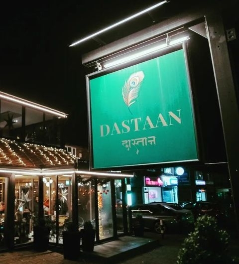 Illuminated signboard of Dastaan with a peacock feather design at night, above the entrance of a well-lit establishment with outdoor seating.