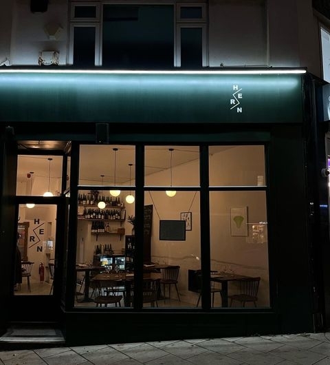 Exterior view of Hern at night, showcasing the illuminated signage above the entrance, with visible interior lighting revealing a cozy dining area with tables and wall art.