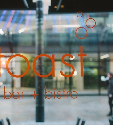 View through a glass window with the words "Toast Bar + Bistro" in orange and white, with a blurred street scene and a pedestrian in the background.
