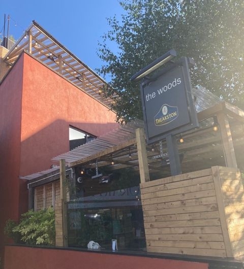 Exterior view of The Woods restaurant with a wooden sign, sunny sky, and partial view of outdoor seating area.