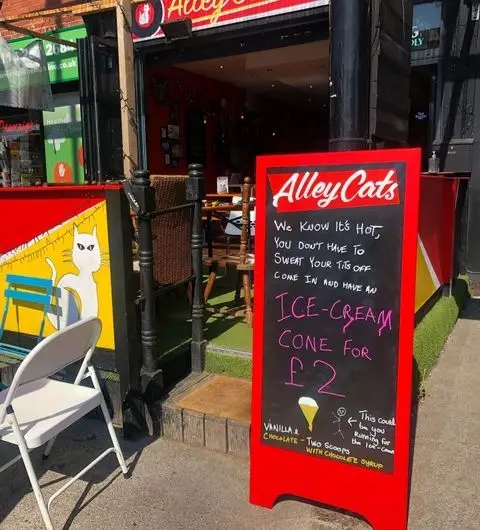 Exterior view of Alley Cats Cafe Bar & Music Emporium with a red sidewalk sign advertising ice-cream cones for £2, featuring a cartoon cat, against a sunny backdrop with outdoor seating.