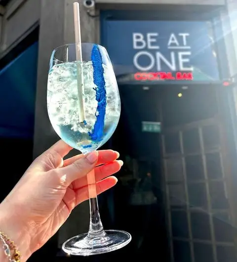 Person holding a blue cocktail in front of Be At One – Boar Lane cocktail bar entrance.