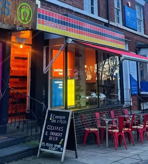 Exterior view of Canteen, featuring a colorful facade with a striped awning, red outdoor seating, and a chalkboard sign advertising cocktails, draught beer, and pizza.