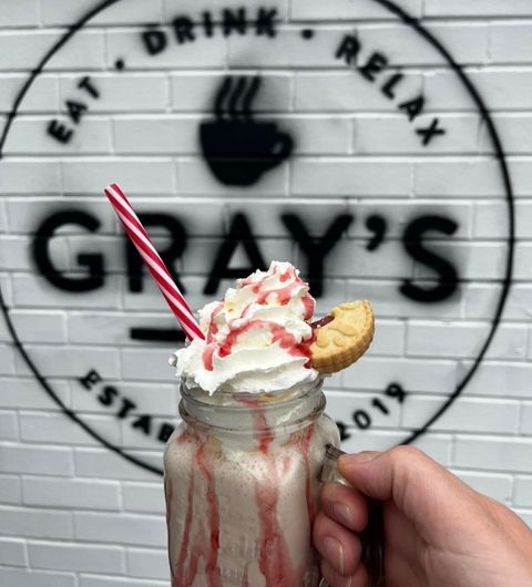 Hand holding a strawberry milkshake topped with whipped cream and a cookie in front of the Gray's logo wall with the words "Eat. Drink. Relax." visible.