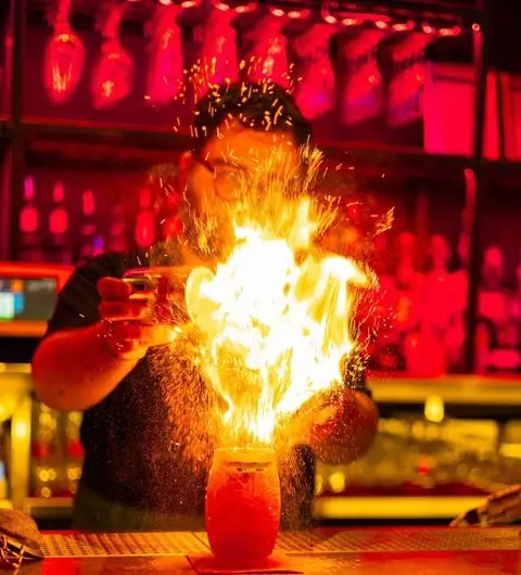 Bartender performing a fire-blowing trick over a cocktail at MOJO bar with a vibrant red backdrop.