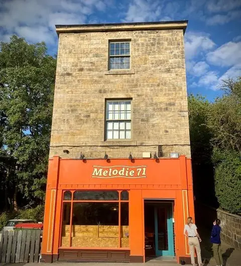 Alt text: "Melodie 71, a quaint three-story building with a distinctive orange storefront and a stone facade, under a clear blue sky with two people walking by."