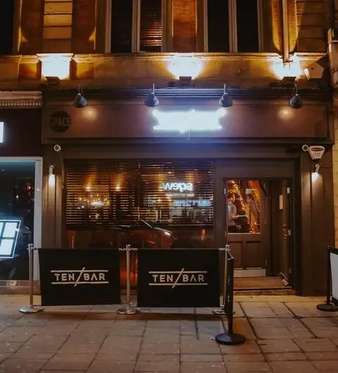 Exterior night view of Ten Bar with illuminated signage, outdoor barriers featuring the bar's name, and warm lighting visible through the front windows.