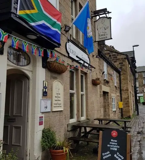 Exterior view of The Horse & Farrier, a traditional pub with hanging flower baskets, colorful bunting, and a variety of flags, including a South African flag, on a cloudy day.