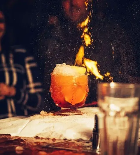 Flaming cocktail at Tiki Hideaway, with orange liquid, ice on top, and a fire burst above the glass on a bar counter.