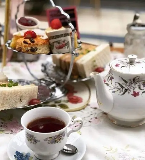 Alt text: "Elegant afternoon tea setup at Just Grand! Vintage Tearoom with a floral porcelain teapot, a cup of tea, and a tiered serving stand filled with sandwiches and cakes."