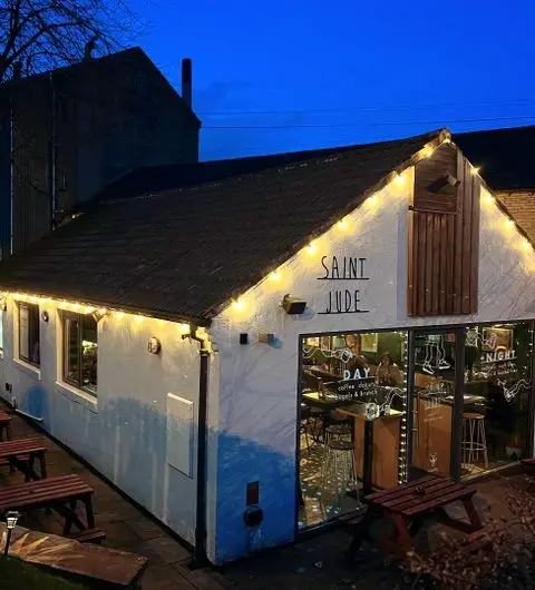 Exterior view of Saint Jude at dusk with warm lighting, featuring the building's white facade, string lights, and outdoor seating area.