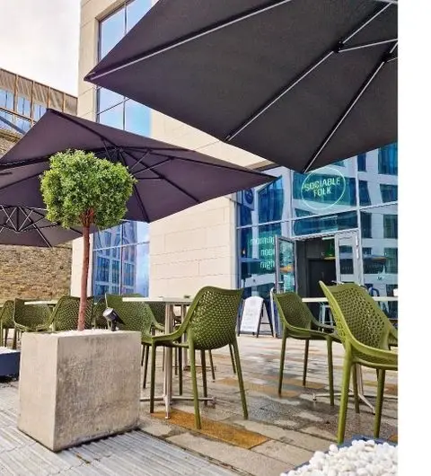 Outdoor seating area at Sociable Folk with green chairs and large umbrellas, a potted tree, and a view of the cafe's entrance.