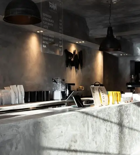 Interior of Raw Coffee shop with a modern, industrial design featuring a concrete counter, black pendant lights, and a menu board displaying coffee options. Bags of coffee and a coffee machine are visible on the counter.