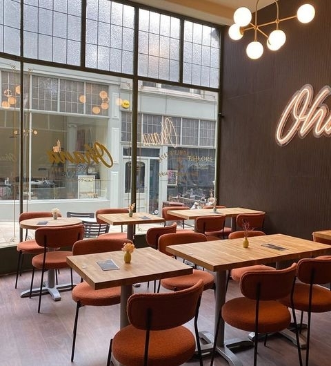 Interior of OHANA cafe with wooden tables, terracotta chairs, and a stylish 'Ohana' neon sign on the wall, large windows provide a view of the street outside.