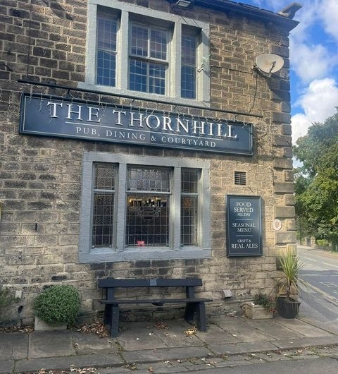 Exterior view of The Thornhill, featuring a stone building with a black sign, bench seating, and a menu board advertising food, seasonal dishes, and real ales.