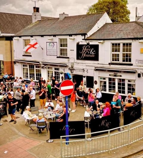 Alt text: A lively outdoor scene at The Hare and Hounds – Rothwell, featuring a crowd of people enjoying drinks and food at tables. The pub's exterior is visible, with the pub sign and an English flag displayed. A red no entry sign is in the foreground.