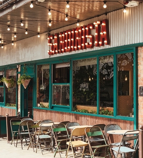 Colorful outdoor seating with metal chairs, string lights above, and a vibrant sign reading "Sandinista" on a rustic facade.