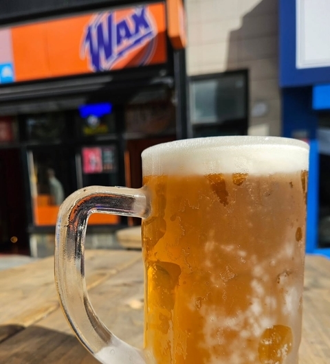 Chilled mug of beer with frothy head in foreground, Wax Bar's vibrant orange sign visible in the background.