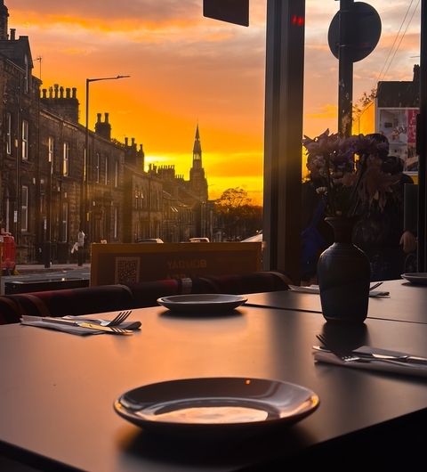 Warm sunset illuminates a cozy dining table at Bombay 2 Goa, with a vase of flowers and cityscape visible outside.
