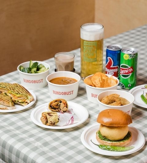 Assorted Indian street food and drinks on a checkered tablecloth at Bundobust, including a sandwich, a burger, bowls of curry, snacks, a pint of beer, and canned beverages.