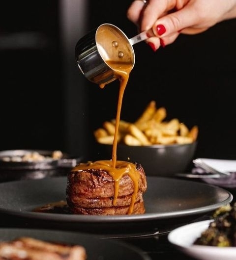 Succulent steak being drizzled with rich, golden sauce at Gaucho, with a side of crispy fries in the background.