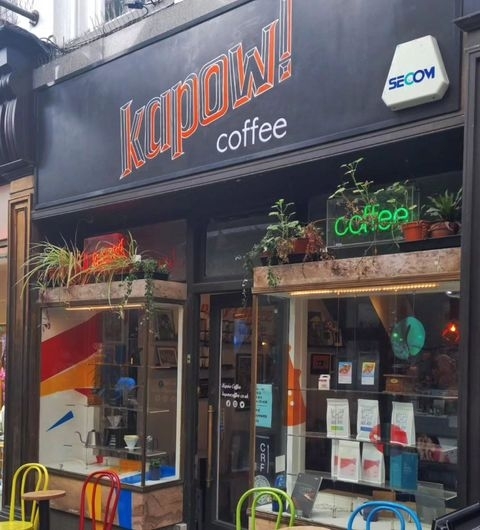Exterior view of Kapow Coffee shop in Thorntons Arcade with a distinctive orange and blue sign, glass front, and potted plants on the window sills.