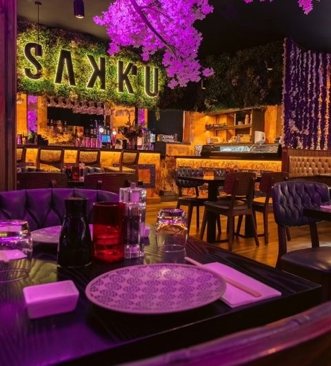 Interior of Sakku restaurant with vibrant purple lighting, decorative cherry blossoms, and a stylish bar area visible in the background.