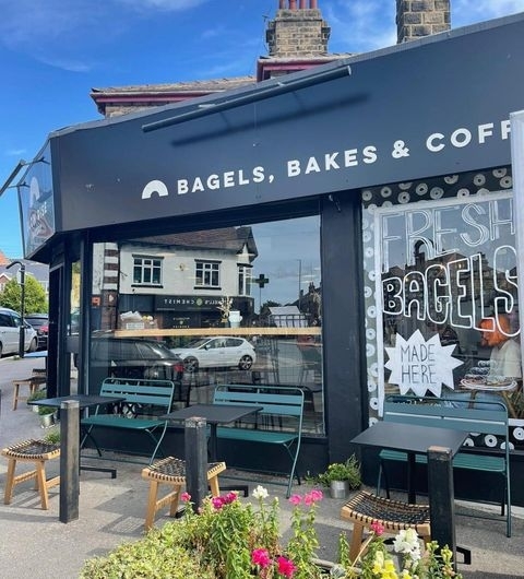 Exterior view of Slow Rise Bakery featuring a black storefront with signage reading 'Bagels, Bakes & Coffee', outdoor seating with green tables and chairs, and a window display advertising 'Fresh Bagels Made Here'. Flowers in the foreground add a welcoming touch.