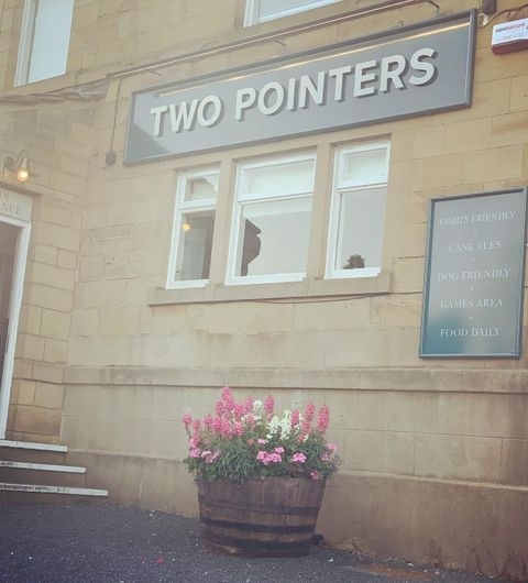 Exterior view of Two Pointers showing the establishment's sign, a broken window, and a signboard with text 'Family Friendly, Cask Ales, Dog Friendly, Games Area, Food Daily' next to a large flower pot with pink blooms.