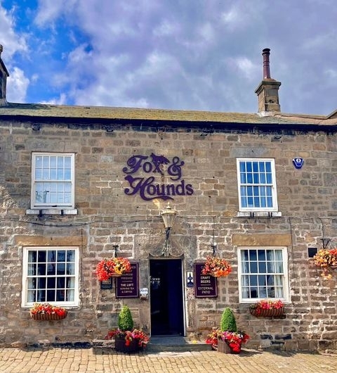 Exterior view of the Fox & Hounds, a traditional stone building with a purple sign, adorned with vibrant flower baskets under a blue sky with scattered clouds.