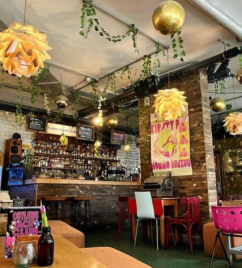 Interior of Outlaws Yacht Club with eclectic decor, featuring a bar lined with various bottles, hanging green plants, and unique lighting fixtures. There are assorted chairs and tables for seating, and a poster on the brick wall adds a pop of color to the space.