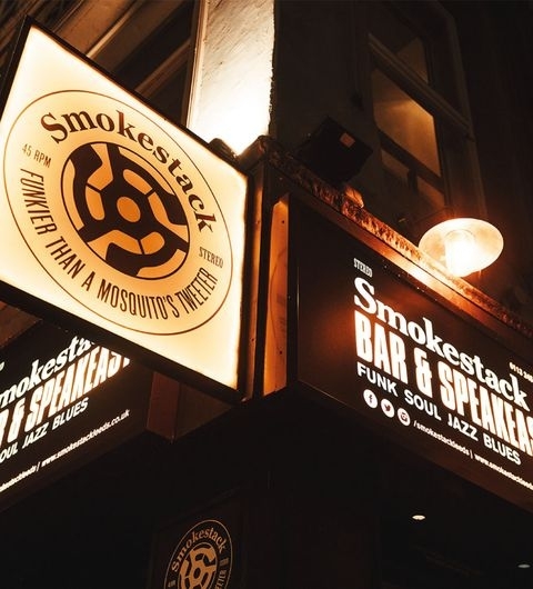 Illuminated signage for Smokestack, featuring the logo and text 'Bar & Speakeasy' with musical genres 'Funk & Soul Jazz Blues' advertised, against a dark evening backdrop.