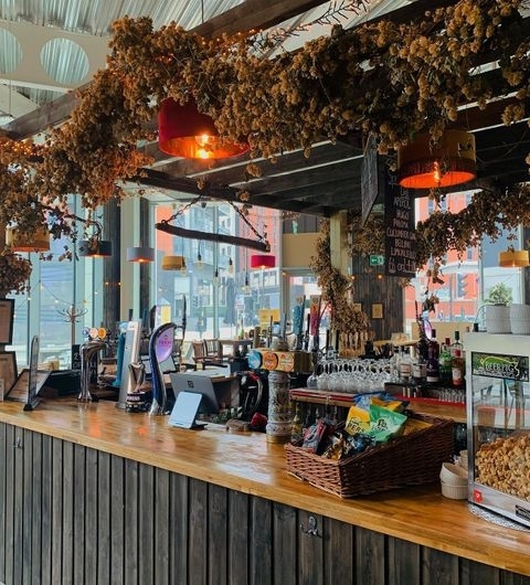 Interior of Hoist House showing a cozy bar area with hanging dried hops, wooden counter, beer taps, pendant lights, and a view of the street outside.