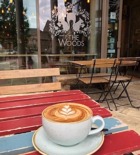 A freshly brewed cappuccino with latte art on a multicolored wooden table at Out of the Woods café, with outdoor seating visible through the glass window.