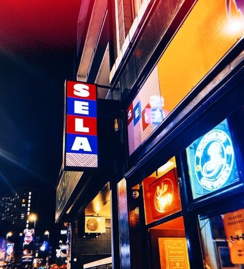 Exterior night view of Sela Bar with illuminated signage and vibrant window displays.