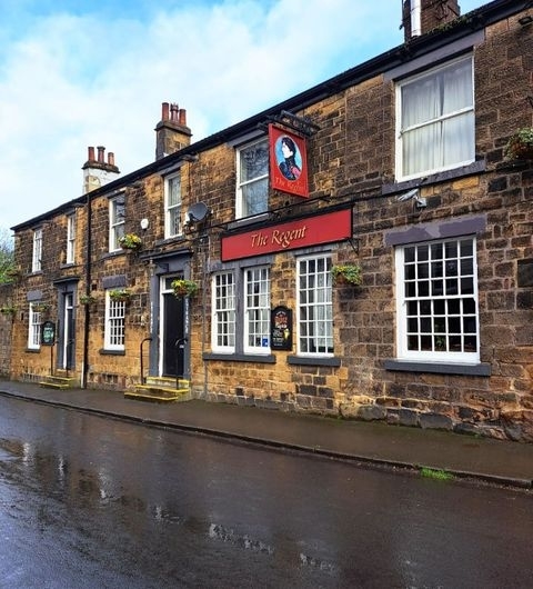 Regent Traditional Pub Architecture Wet Street Exterior view of The Regent, a traditional stone-built pub with a red signboard, on a wet street reflecting the cloudy sky above.