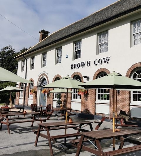 Exterior view of The Brown Cow pub with outdoor seating and green umbrellas on a sunny day.