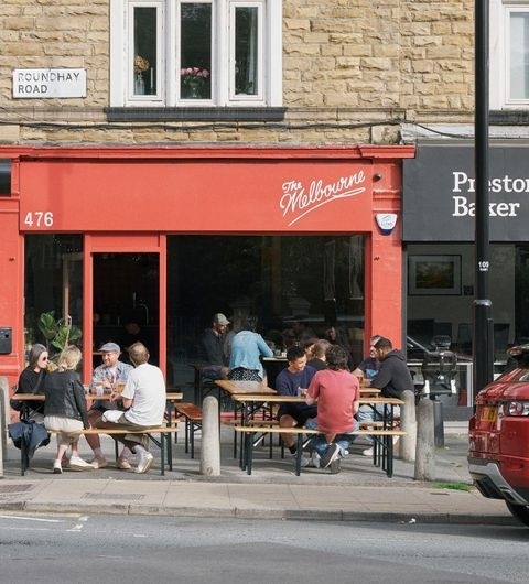 People enjoying outdoor seating at The Melbourne cafe with a vibrant red storefront on a sunny day.