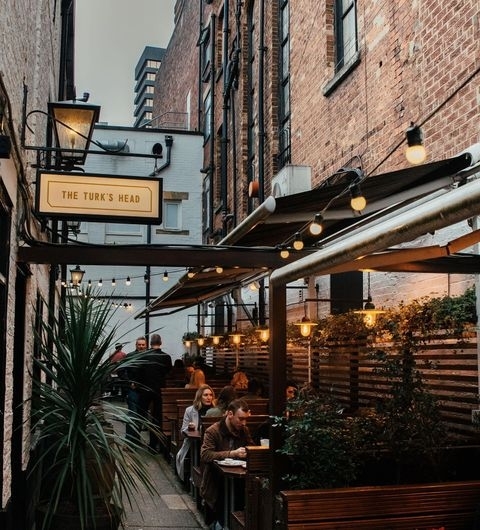 Outdoor seating area of The Turk's Head with patrons dining under string lights, surrounded by brick buildings and greenery.