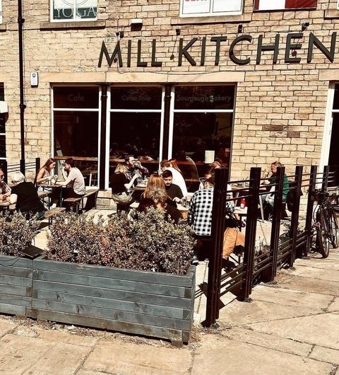Outdoor seating at Mill Kitchen & Bakery with patrons enjoying meals under sunny skies, surrounded by greenery and parked bicycles.