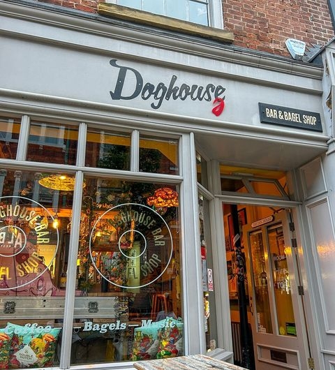 Exterior view of Doghouse Bar & Bagel Shop with illuminated signage, glass windows showcasing interior lights and decorations, and an open entrance door.