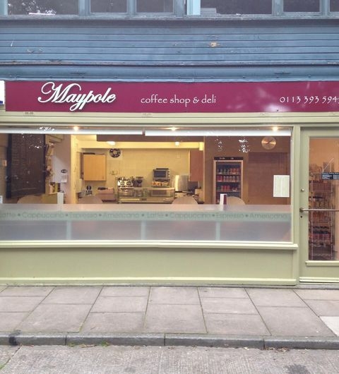 Exterior view of Maypole Coffee Shop & Deli with a maroon and white signboard, large glass windows showcasing the interior with a coffee machine and shelves, and a pastel green storefront.