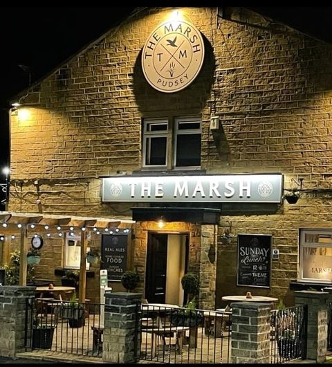 Exterior view of The Marsh – Pudsey at night, showcasing its illuminated signage and warm lighting, with a stone facade and welcoming entrance.