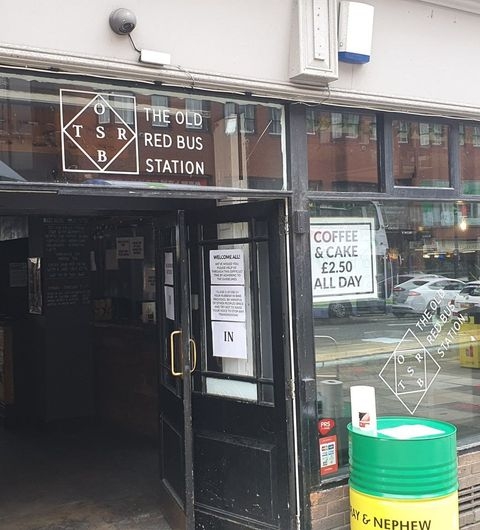 Entrance to The Old Red Bus Station with a sign above the door, offering coffee and cake, and a view of the street reflected in the glass window.