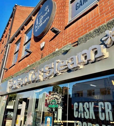 The Head of Steam Headingley, a vibrant pub with a modern exterior, features a prominent sign showcasing its name. The building has a mix of brick and glass, with large windows allowing for a view inside. The entrance is welcoming, with decorative elements and outdoor seating visible.