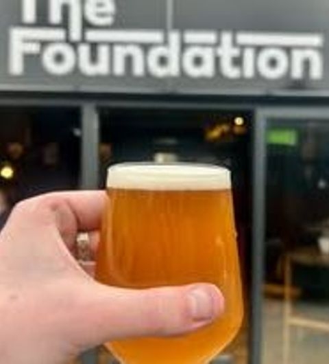 A hand holding a glass of amber beer in front of the entrance to The Foundation, showcasing the establishment's sign in the background.